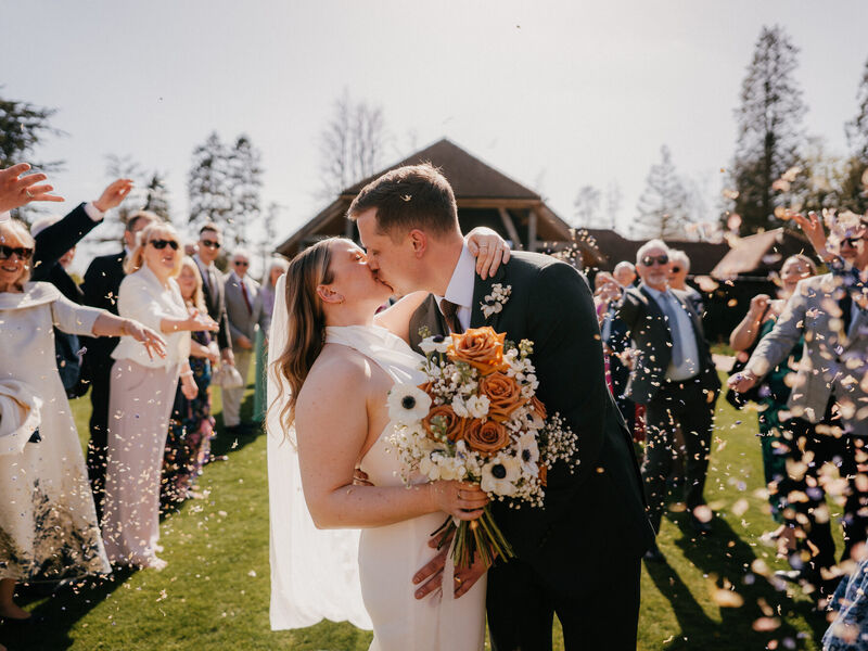 Sophie and Grant kissing beneath confetti with a toffee rose and white anemone bouquet
