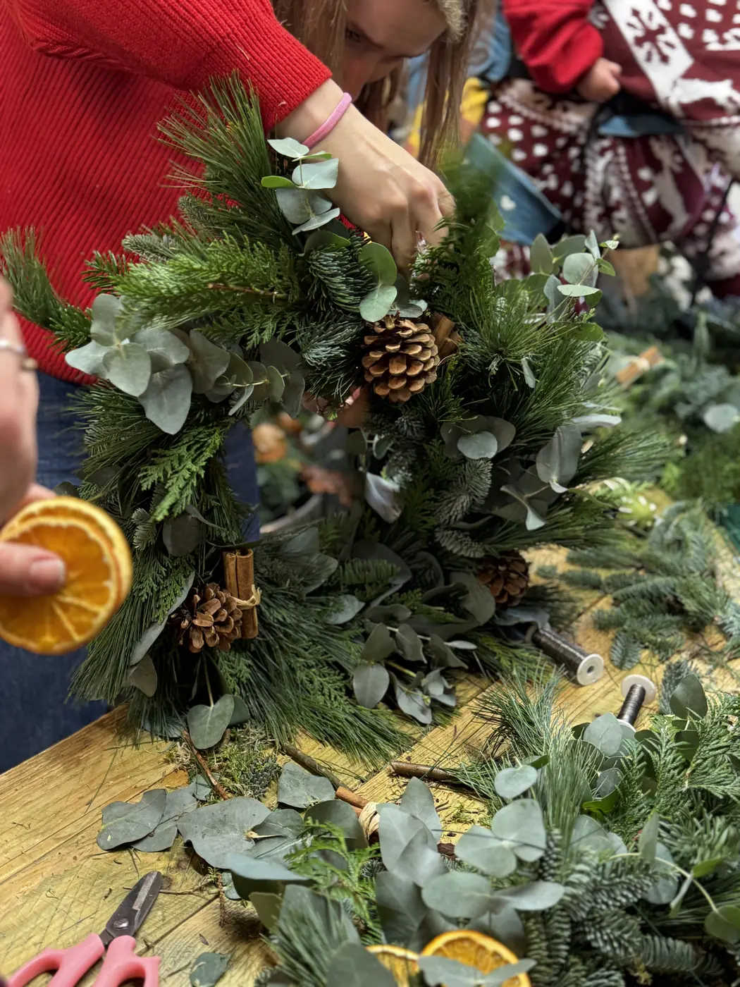 Hands adding evergreen foliage and pine cones to a natural Christmas wreath